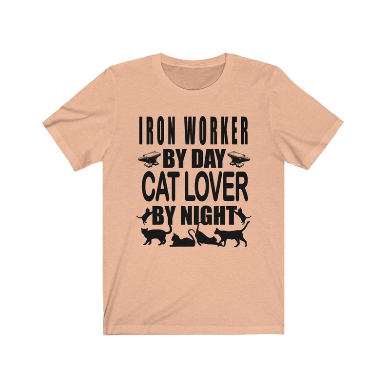 May include: Peach colored t-shirt with black text that reads "Iron Worker By Day Cat Lover By Night". The shirt has a black silhouette of an anvil and four cats.