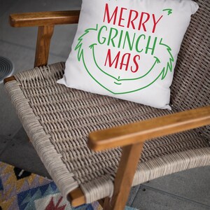 May include: White throw pillow with green and red text that reads "Merry Grinch Mas" in a whimsical font. The pillow is sitting on a woven chair with a wooden frame.