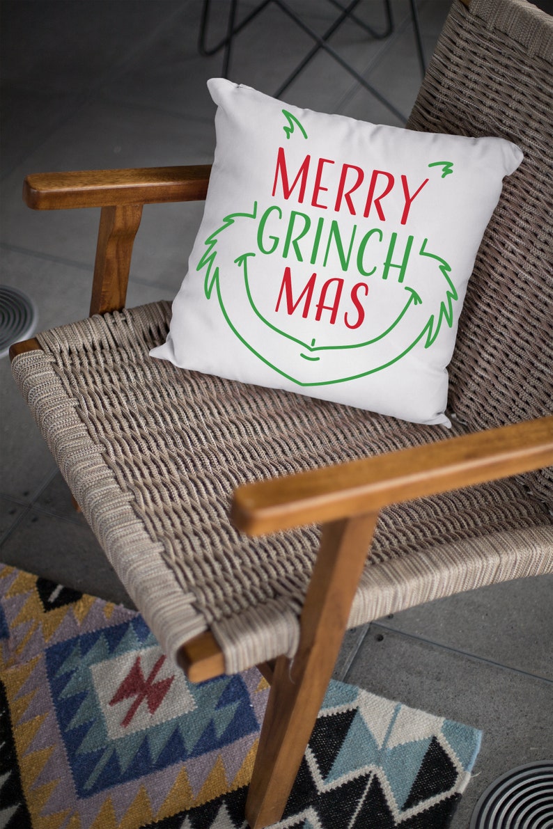May include: White throw pillow with green and red text that reads "Merry Grinch Mas" in a whimsical font. The pillow is sitting on a woven chair with a wooden frame.
