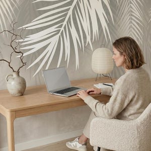 May include: A wall mural featuring large, white palm leaf silhouettes against a soft, neutral-toned background. The design covers the wall behind a light-colored wooden desk, a ceramic vase with bare branches, a laptop, and a paper lantern lamp.