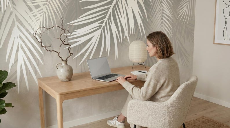May include: A wall mural featuring large, white palm leaf silhouettes against a soft, neutral-toned background. The design covers the wall behind a light-colored wooden desk, a ceramic vase with bare branches, a laptop, and a paper lantern lamp.