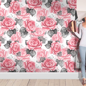Puede incluir: Una mujer est&aacute; colocando papel tapiz floral rosa y gris con un dise&ntilde;o de acuarela en una pared. El papel tapiz presenta un patr&oacute;n repetitivo de rosas rosas y hojas grises.