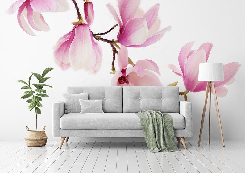 May include: A white couch with a green throw blanket sits in a room with a white wall featuring a pink magnolia flower mural. A small potted plant and a floor lamp are also in the room.