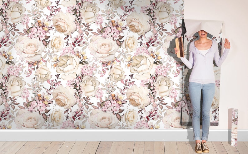 K&ouml;nnte beinhalten: Eine Frau h&auml;ngt eine Tapete mit einem Blumenmuster mit wei&szlig;en Rosen, rosa Bl&uuml;ten und braunen Bl&auml;ttern auf wei&szlig;em Hintergrund.