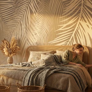 May include: A bedroom featuring a wall mural with large, light-colored palm leaf silhouettes against a warm beige background. The bed has a woven rattan headboard, layered neutral-toned bedding, and a textured throw blanket. Two woven baskets sit at the foot of the bed, and a dried floral arrangement in a vase is placed on the bedside table.
