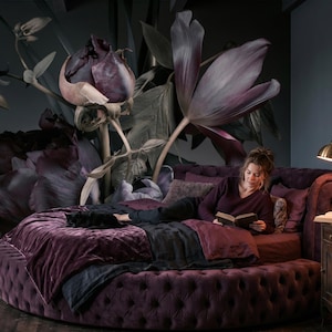 May include: A round, tufted, deep purple velvet bed frame with matching bedding, featuring a large-scale floral wall mural in shades of dark purple and black. A brass desk lamp sits on a wooden nightstand next to the bed.