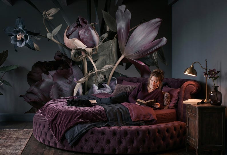 May include: A round, tufted, deep purple velvet bed frame with matching bedding, featuring a large-scale floral wall mural in shades of dark purple and black. A brass desk lamp sits on a wooden nightstand next to the bed.