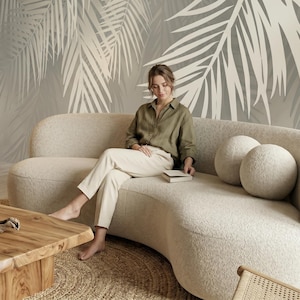 May include: A curved, cream-colored boucl&eacute; sofa with two matching round throw pillows. The sofa features a rounded backrest and a deep seat, providing a soft, textured appearance. It is positioned in a room with a large palm leaf wall mural.