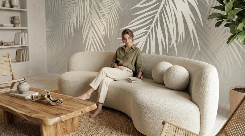May include: A curved, cream-colored boucl&eacute; sofa with two matching round throw pillows. The sofa features a rounded backrest and a deep seat, providing a soft, textured appearance. It is positioned in a room with a large palm leaf wall mural.