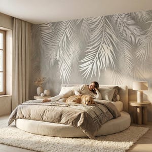 May include: A large round bed with beige bedding and a golden retriever lying on top. The wall behind the bed features a large-scale mural of white palm fronds on a light gray background. A cylindrical lamp sits on a round wooden side table.