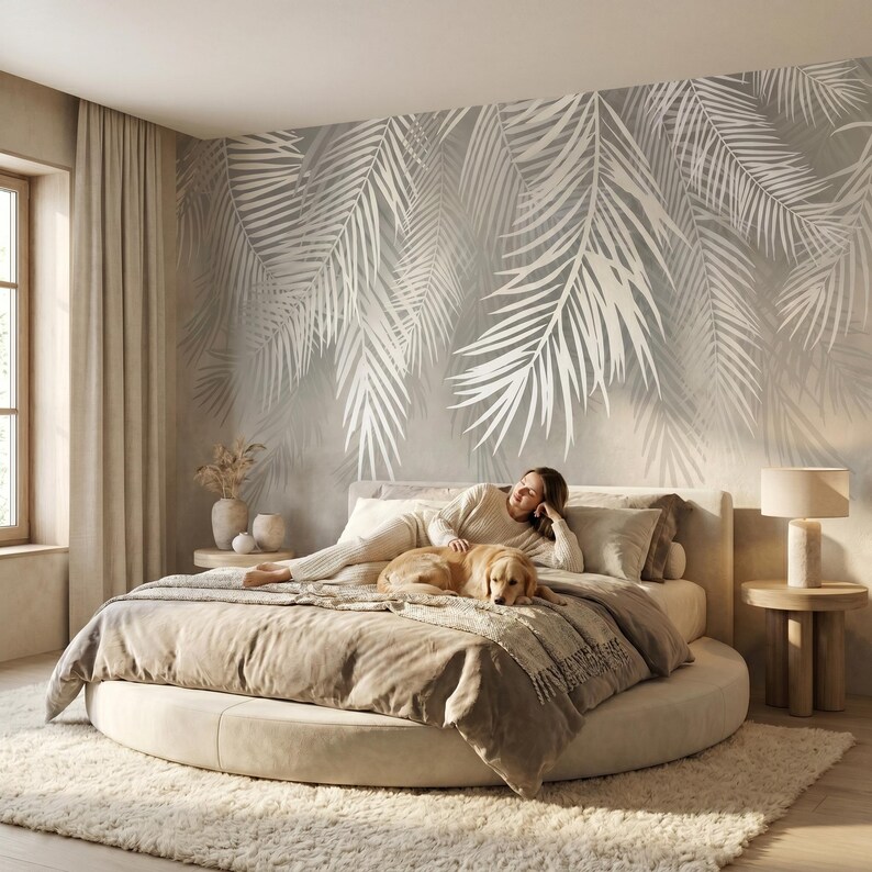 May include: A large round bed with beige bedding and a golden retriever lying on top. The wall behind the bed features a large-scale mural of white palm fronds on a light gray background. A cylindrical lamp sits on a round wooden side table.