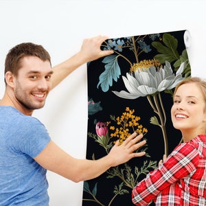 May include: A couple are hanging up a black and white floral wallpaper with a large white flower in the centre. The wallpaper has a dark background with green leaves and other flowers in shades of pink, yellow, and blue.