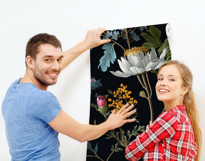May include: A couple are hanging up a black and white floral wallpaper with a large white flower in the centre. The wallpaper has a dark background with green leaves and other flowers in shades of pink, yellow, and blue.