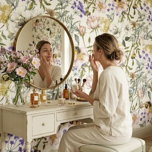 May include: A round gold-framed mirror sits on a white wooden vanity table with two drawers. A clear glass vase holds pink flowers. Several skincare bottles and jars are arranged on the surface. The background features floral wallpaper.