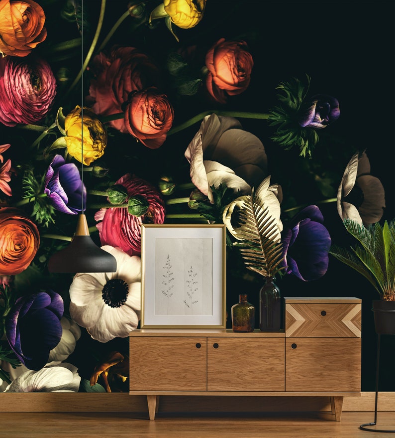 May include: A dark wood cabinet with two drawers and two doors, featuring a floral wallpaper with a variety of colourful flowers, including red, yellow, purple, and white. A gold framed picture hangs above the cabinet, and a gold leaf plant sits on the cabinet.