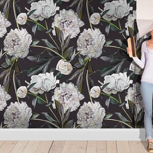 May include: A woman is standing in front of a wall covered in white peony floral wallpaper. The wallpaper has a dark background and features white peonies with green leaves. The woman is holding a roll of wallpaper and is smiling.
