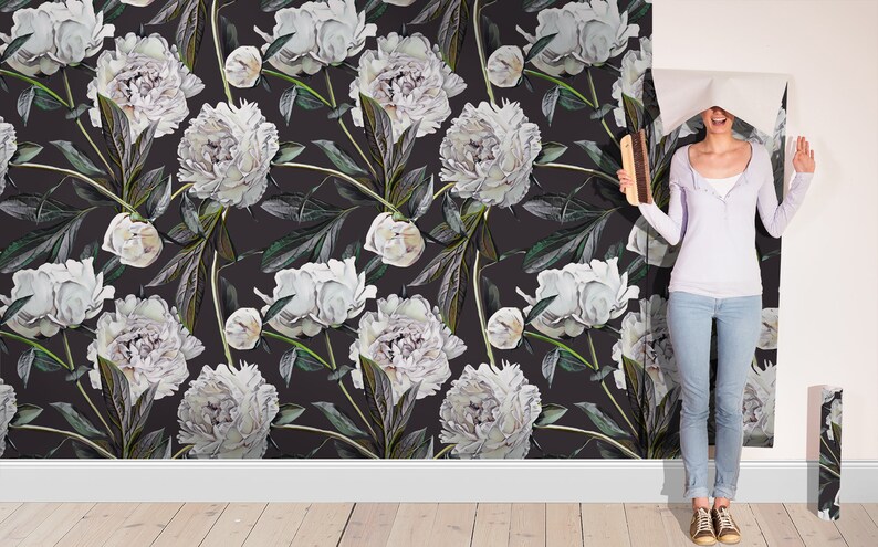 May include: A woman is standing in front of a wall covered in white peony floral wallpaper. The wallpaper has a dark background and features white peonies with green leaves. The woman is holding a roll of wallpaper and is smiling.