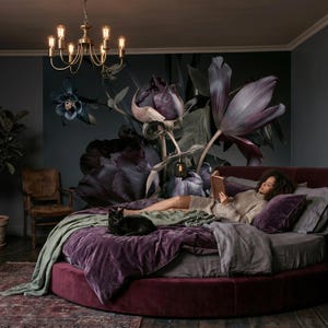 May include: A round, deep burgundy velvet bed frame with matching plush bedding, a sage green throw blanket, and a black cat resting on top. The bedroom features dark walls with a large floral mural, wooden furniture, and a brass chandelier.