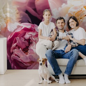 以下が含まれることがあります： 子供を含む3人家族が、大きな花の壁画のある壁の前にある白いソファに座っています。壁画にはピンクと白の牡丹が描かれています。小さな白と茶色の犬がソファの前に座っています。