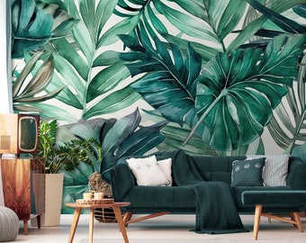 May include: A watercolor-style mural featuring large, lush green tropical leaves. The leaves vary in shades of green, creating a vibrant and naturalistic design. The mural is installed behind a dark green sofa with white pillows, a wooden side table, and a vintage-style cabinet.