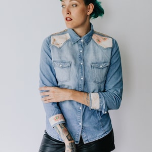 May include: Light blue denim shirt with decorative patches on the shoulders and cuffs. The shirt has a button-down front, two chest pockets, and long sleeves. The patches feature embroidered designs with earth-tone colors. The model is wearing black shorts and sheer tights.