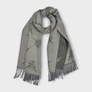 May include: A gray scarf with a repeating pattern of Highland cow illustrations. The scarf has a fringed edge and a two-tone design, with a darker gray panel on one side. The scarf is made of a soft material.