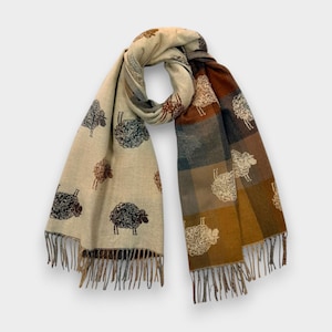 May include: A long scarf featuring a two-tone design. One side is a light beige with dark brown sheep illustrations, while the other side displays a brown and blue plaid pattern with white sheep motifs. The ends are finished with fringe detailing.