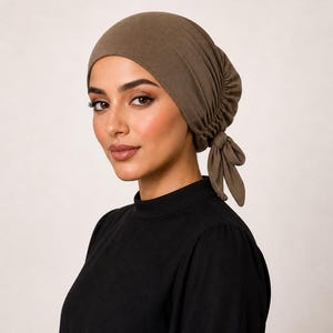 May include: A brown head covering with a gathered design and a tie closure. The head covering is made of a soft fabric and is worn on the head. The person in the image is wearing a black top.