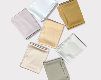 May include: Six satin hijabs in different colors: white, light gray, beige, light brown, gold, and olive green. The hijabs are folded and arranged in a circle on a white background. The text "SATIN HIJABS" is written in gold letters below the hijabs.