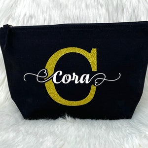 Könnte beinhalten: Schwarze Canvas-Kosmetiktasche mit einem goldenen Glitzerbuchstaben C und dem Namen Cora in weißer Kursivschrift.
