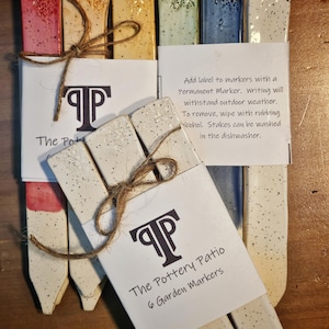 May include: A collection of ceramic garden markers in various colors, including red, yellow, green, and blue, with speckled white bases. Each marker is tied with twine and packaged with a card that reads "The Pottery Patio 6 Garden Markers."