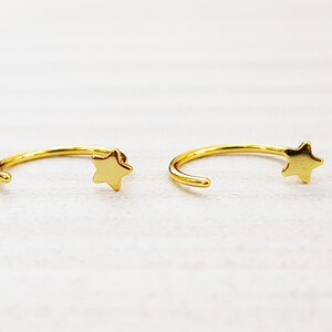 Puede incluir: Un par de pendientes de aro de oro con un dije de estrella en cada aro.