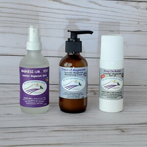 May include: Three bottles of lavender magnesium products from Big Sky Lavender Farms. The products are a spray, a cream, and a roll-on. The spray bottle is clear with a white label. The cream bottle is brown glass with a white label. The roll-on bottle is white with a blue label.