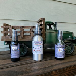 K&ouml;nnte beinhalten: Drei kleine Glasspr&uuml;hflaschen mit Etiketten. Die Flaschen stehen auf einer Holzoberfl&auml;che vor einem Spielzeuglaster. Die Etiketten lauten "Calm Your Down It's a Guy Thing! Sky Lavender Farms", "De-Buggin Me Mosquito Repellent Bugger-Off Strength Sky Lavender Farms Deet Free", und "Man-Cave Magnesium It's a Guy Thing! Sky Lavender Farms".