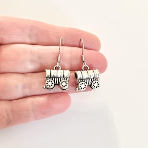 May include: Silver dangle earrings featuring a wagon design. The wagon is detailed with wheels and a roof.