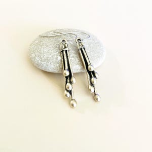 May include: Silver dangle earrings with a textured, abstract design. The earrings feature multiple small, round beads hanging from a long, thin bar.