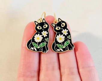 May include: A pair of black cat earrings with gold-colored hooks. Each cat is decorated with white daisy flowers and green leaves. The earrings are held up by a hand.