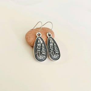 May include: Silver dangle earrings with a teardrop-shaped design featuring a raised image of two pine trees.