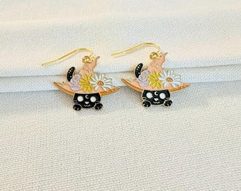 May include: A pair of gold-tone hook earrings featuring a black cat wearing a light peach witch hat adorned with white and yellow daisies and a small purple flower. The cat has a small white crescent moon symbol on its forehead and gold hooks.