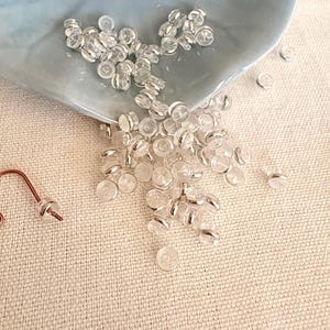 May include: A collection of small, clear, circular earring backs with silver-toned metal accents. These translucent, disc-shaped components are designed to secure earring posts, featuring a central hole for insertion and a smooth, rounded profile.