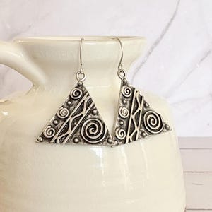 May include: A pair of silver-toned triangle earrings. Each earring features an intricate design of spirals, dots, and lines. The earrings hang from simple ear wires. The earrings are displayed against a white background.