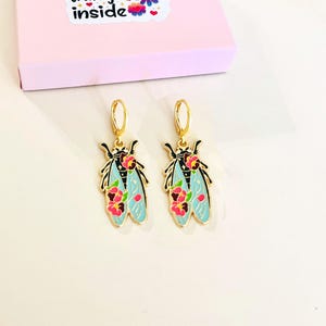 May include: A pair of gold-colored hoop earrings featuring a cicada design. The cicadas have light blue wings with pink floral accents and black outlines. A pink box with the words "inside" is in the background.