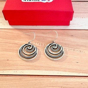May include: A pair of silver-toned earrings with a circular design featuring swirling lines. The earrings are displayed on a wooden surface, with a red box in the background. The box has a sticker that says "things inside".