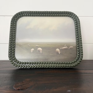 May include: A rectangular picture frame with a beaded, olive green border. The framed image depicts a pastoral scene of sheep grazing in a field under a cloudy sky. The frame is displayed on a dark wooden surface.