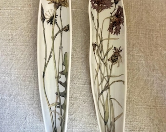 May include: Two elongated, narrow ceramic dishes with a smooth, off-white finish. Each dish features a detailed botanical illustration of wildflowers in muted tones of brown, green, and yellow, extending along the interior length of the piece.