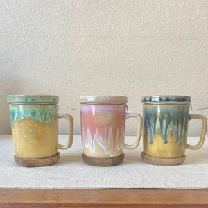 May include: Three ceramic tea mugs with lids and infusers. Each mug has a unique glaze: yellow and turquoise, pink and white, and yellow and blue. The mugs sit on wooden coasters and have matching handles.