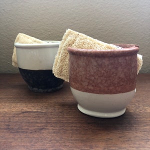 May include: Two ceramic soap dishes with a white and brown glaze. The dishes are filled with natural loofah sponges.