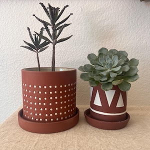 May include: Two terracotta plant pots with white geometric designs. The pot on the left has a pattern of small white dots. The pot on the right has a pattern of white triangles. Both pots have matching saucers.