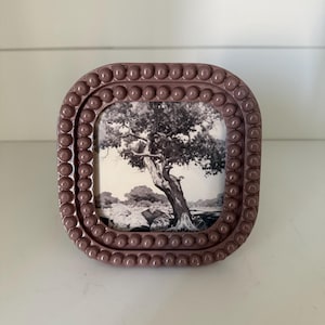 Op de afbeelding: Een mauve vierkante fotolijst met een afgeronde rand en een kralenrand. De lijst bevat een zwart-witfoto van een boom in een landschap. De lijst wordt weergegeven op een wit oppervlak.