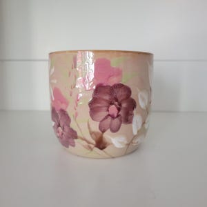 Könnte beinhalten: Ein zylindrischer Keramikbecher mit einer Basis in sanftem Pfirsichton, verziert mit handgemalten Blumenmotiven in dunklen Pflaumen-, leuchtenden Pink- und Weißtönen. Das Design umfasst abstrakte Blattmuster und Pinselstriche auf der Außenseite.